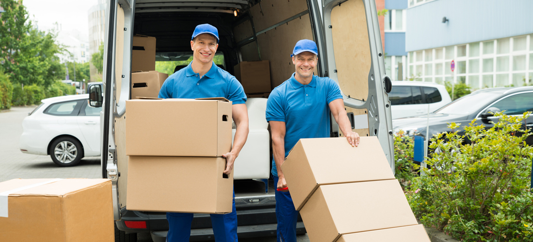 Two Happy Male Workers Loading Stack Of Cardboard Boxes In Truck