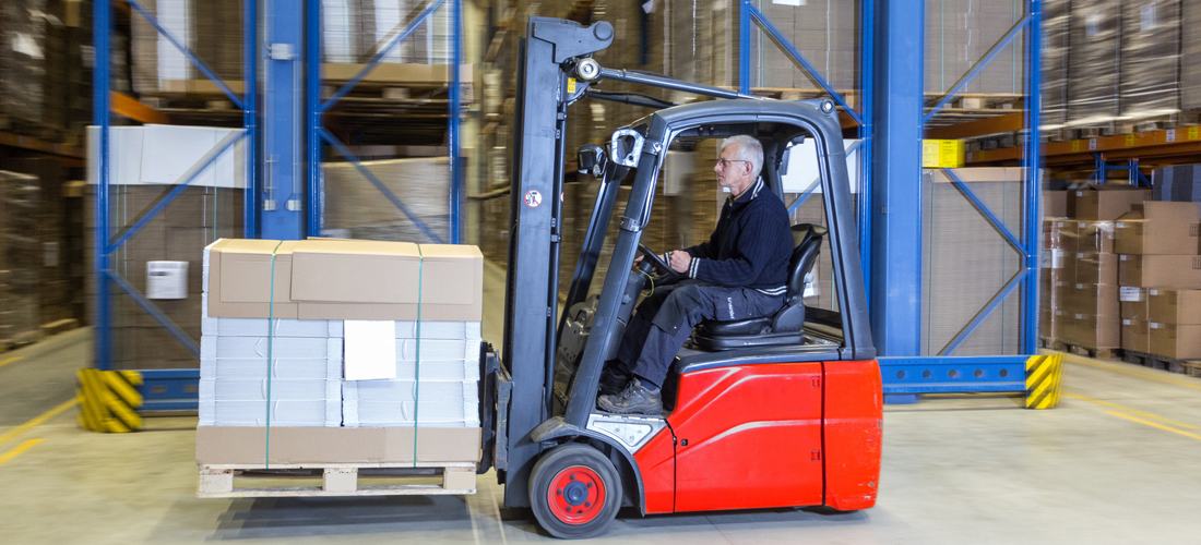 Man is driving a forklift alongside a few rows of storage racks. The experienced driver is transporting a pallet full of cardboard boxes.